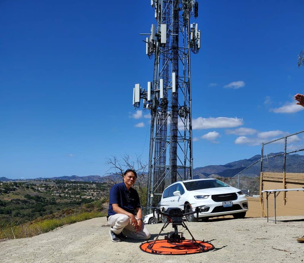 A person kneels beside a drone on a landing pad near a white car, showcasing telecoms innovation with a cell tower and mountains in the background under a blue sky. Engineering for Impact by Apurba Tribedi.