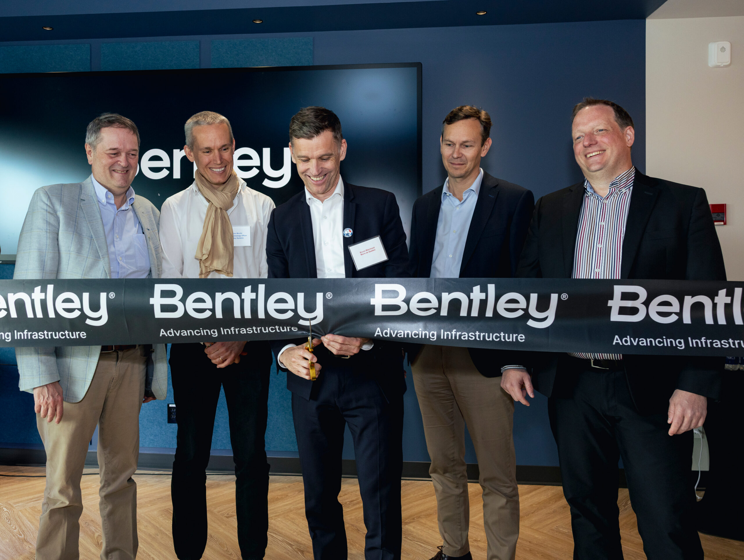 Five men in business attire stand together at a ribbon-cutting event for Bentley, holding a ribbon that reads "Bentley Advancing Infrastructure.