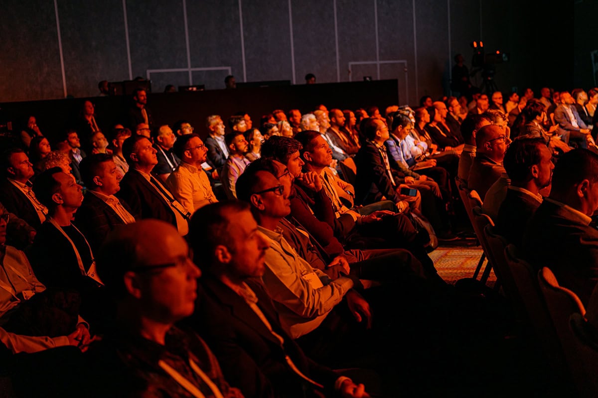 A large audience sits in rows of chairs, attentively watching a presentation or event in a dimly lit conference room.
