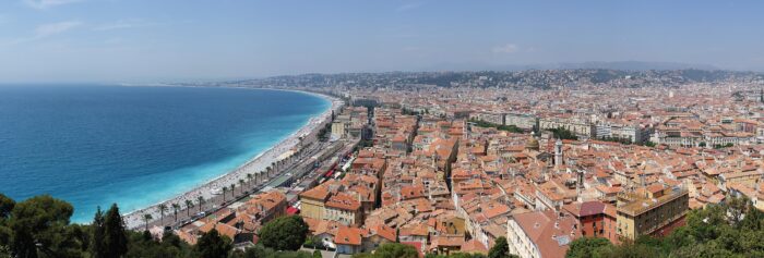 Panoramic view of Nice, France, featuring the Mediterranean Sea, palm-lined Promenade des Anglais, and terracotta rooftops against a clear blue sky.