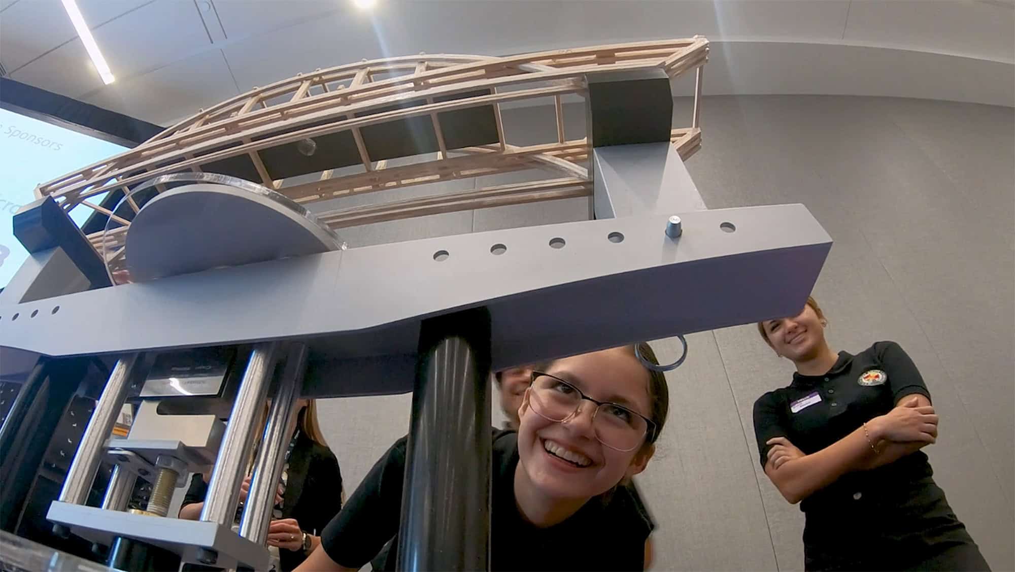 A person smiles underneath a model bridge structure, with others standing in the background observing.