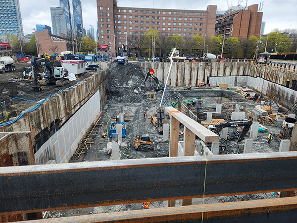 Construction site with excavated foundation, construction materials, machinery, and workers present. Toronto rail protection measures highlight rail safety innovation as city buildings and trees are visible in the background.