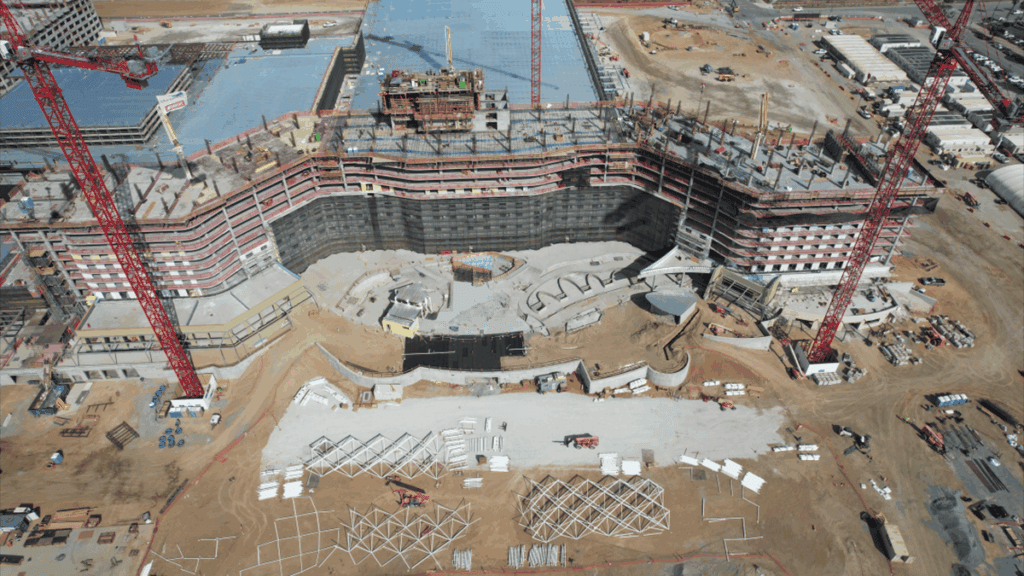 Aerial view of a large construction site with cranes, partially built structures, and building materials spread across sandy ground.
