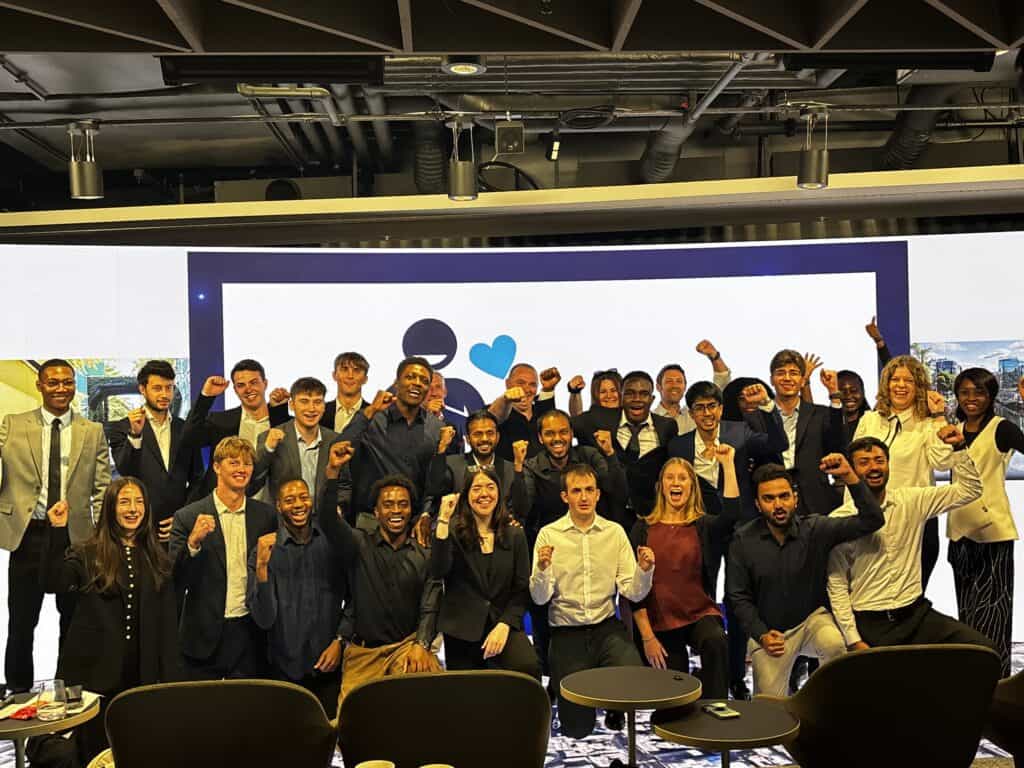 A large group of people in business attire pose together indoors, some with raised fists, in front of a large screen displaying a simple graphic, celebrating their achievements at a global contest.