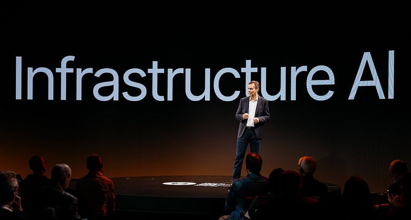 A person stands on stage giving a presentation with the words "Infrastructure AI" displayed in large letters on a screen behind them.