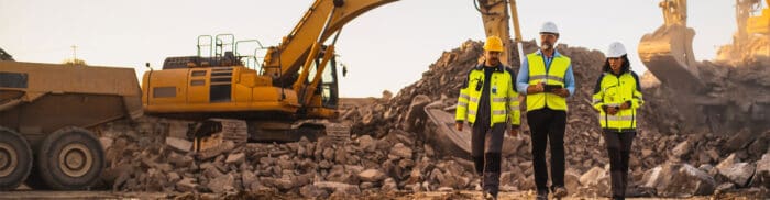 Three construction workers in high-visibility jackets and helmets walk through a rocky construction site with heavy machinery in the background.