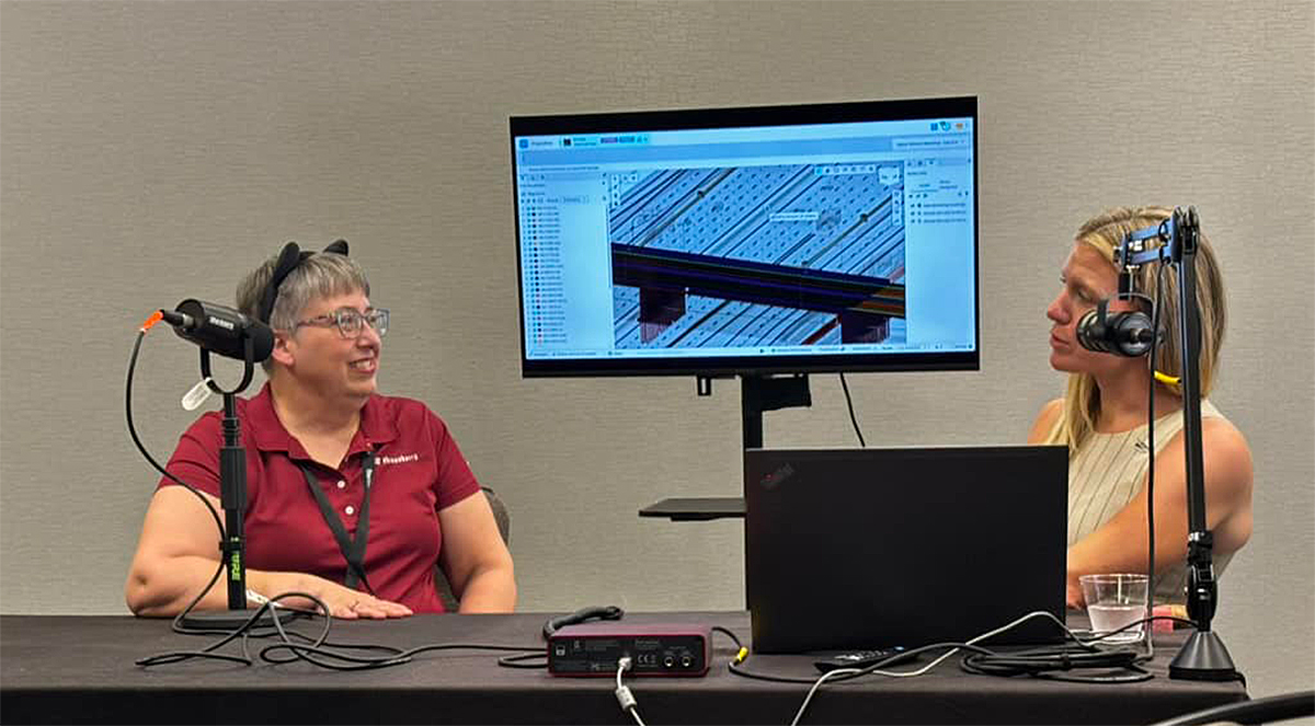 Two women sit at a table with microphones and a laptop, recording a podcast or interview. A monitor behind them displays a technical diagram or blueprint.