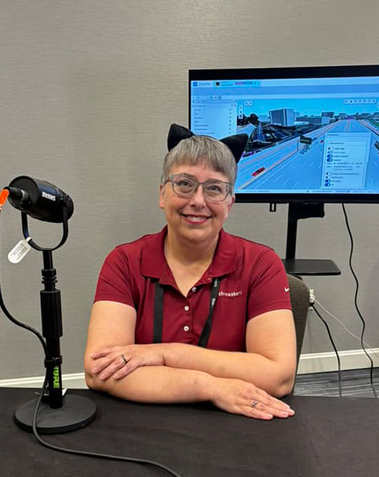 A person in a red polo shirt sits at a table with a microphone and cables, in front of a monitor displaying a software interface.