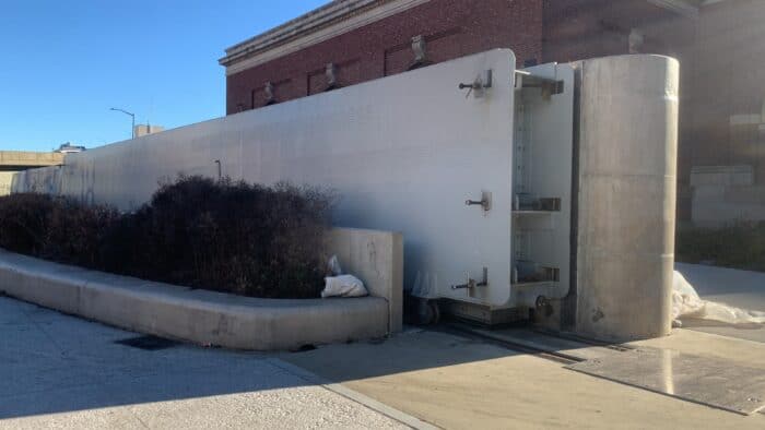 Large metal doors closed across a street entrance next to a brick building, with concrete pillars and bushes nearby.