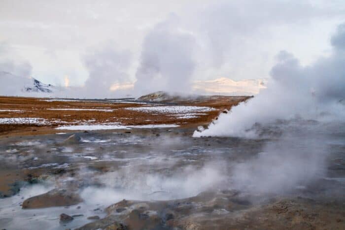 Steam rises from geothermal vents, showcasing the power of geothermal energy in a rocky, snow-dusted landscape under a cloudy sky.