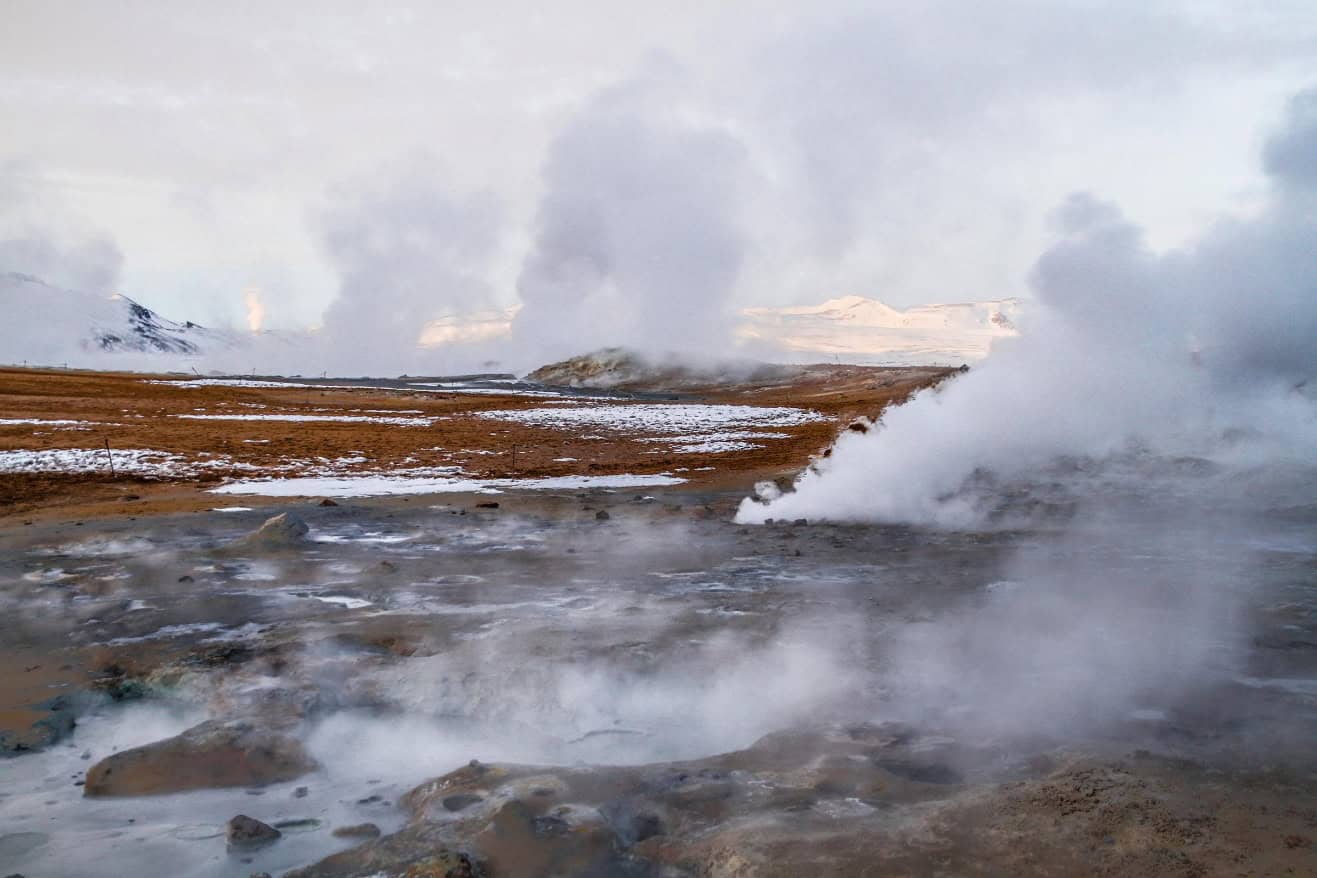 Steam rises from geothermal vents, showcasing the power of geothermal energy in a rocky, snow-dusted landscape under a cloudy sky.