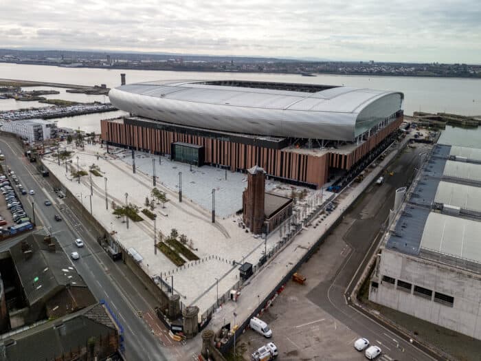 Aerial view of a large, modern stadium with a silver roof near a river, surrounded by parking and roads. Overcast sky and cityscape in the background.