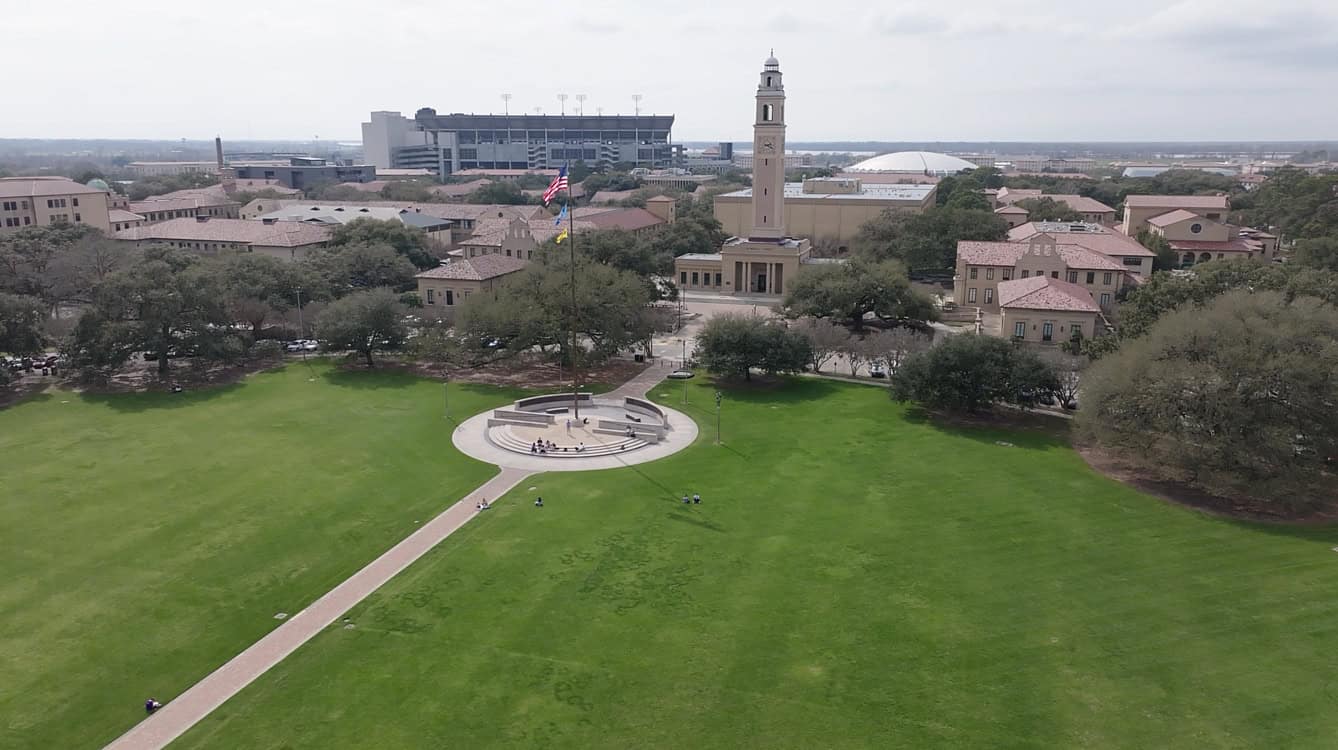 Aerial view of a large grassy field with a flagpole and pathways, surrounded by trees and buildings. A tall clock tower is prominent. A stadium is visible in the background.
