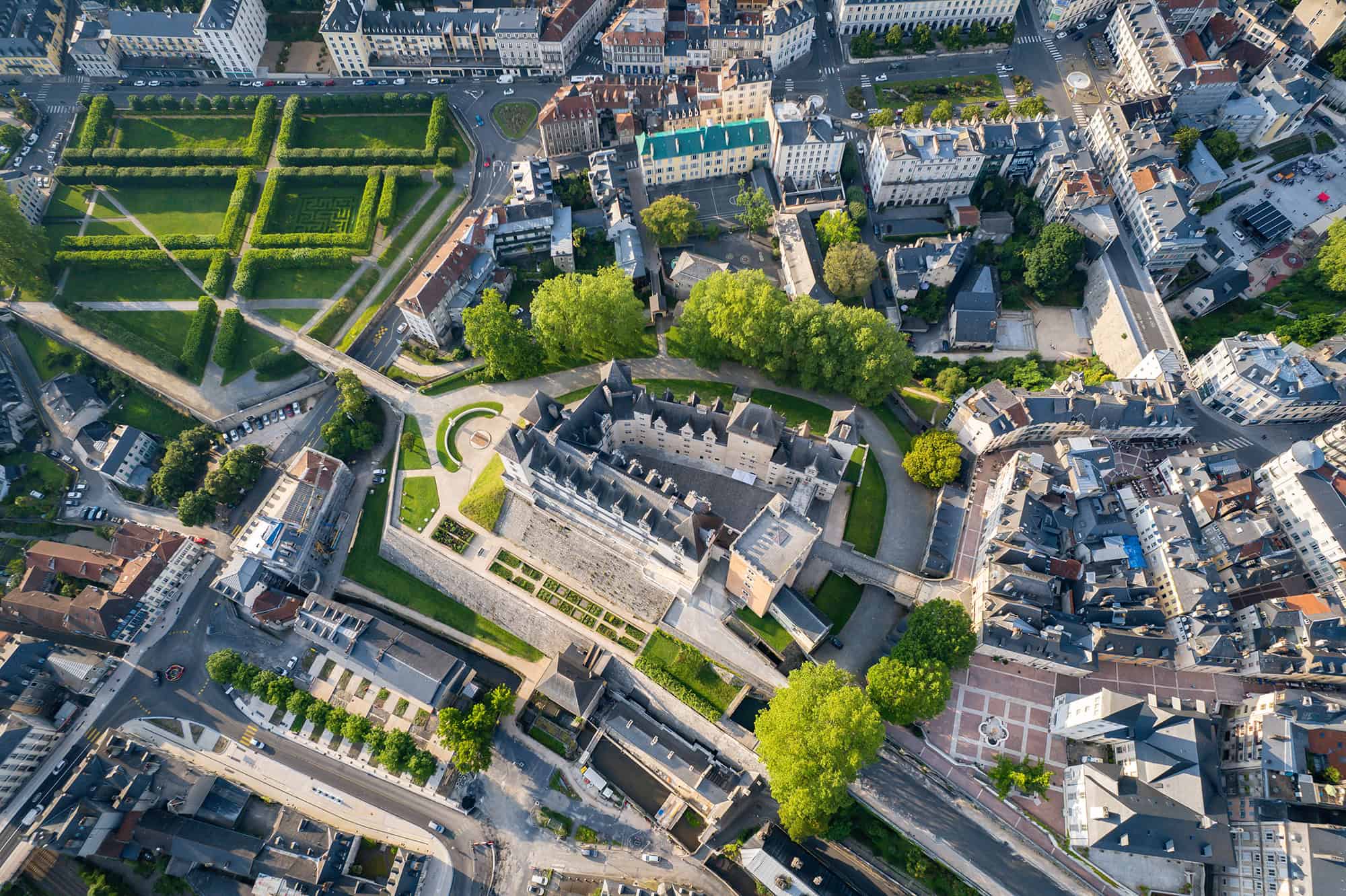 Aerial view of a historic castle complex with surrounding gardens and city buildings, intersected by roads and tree-lined pathways.