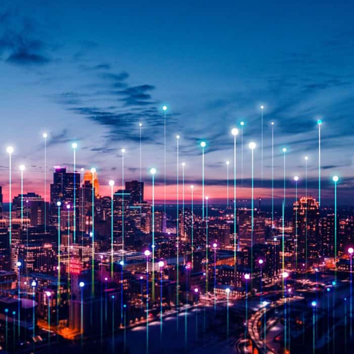 A city skyline at dusk with illuminated colored lines and dots digitally superimposed, representing AI-driven technology or data connectivity shaping urban infrastructure.