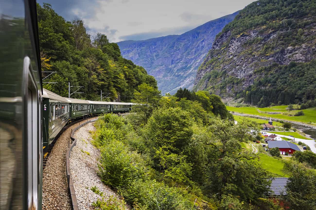 A long passenger train curves through green, mountainous terrain with forested hills and small buildings visible in the valley below, much like the innovative solutions found in the Toronto rail corridor that prioritize both scenic travel and advanced rail protection.