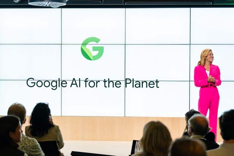 A woman in a bright pink suit stands on stage in front of a screen displaying the text “Google AI for the Planet” to an audience.