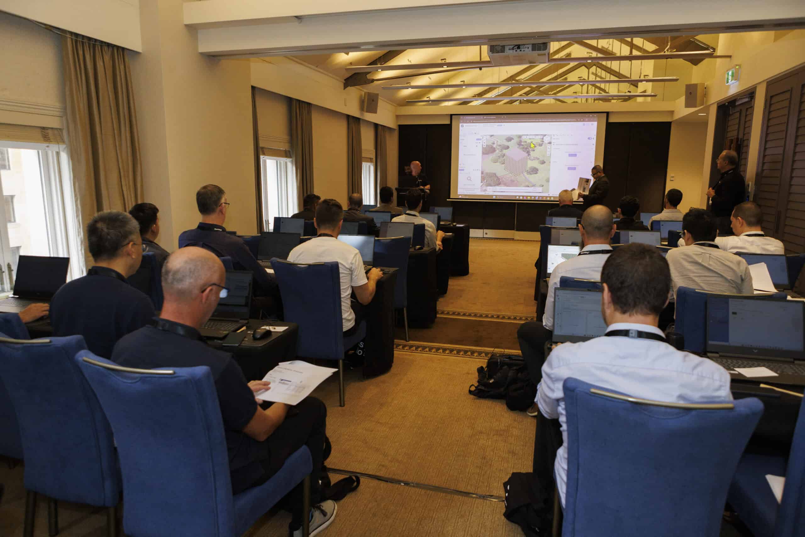 People seated in a conference room with laptops, watching a presenter speak at the front near a screen displaying a map.