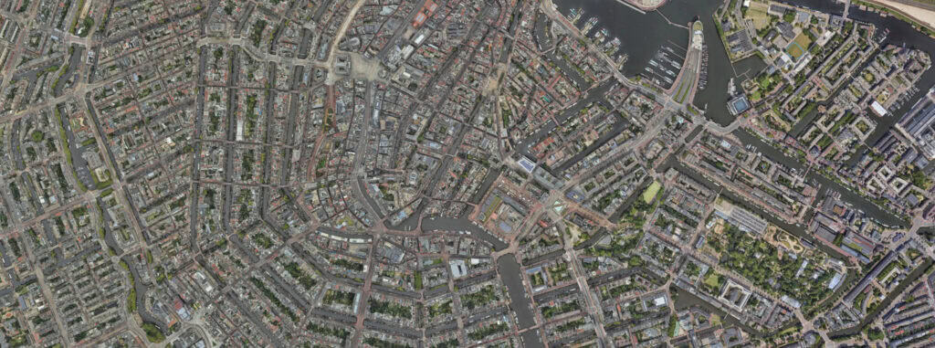Aerial 3D view of a densely built Netherlands city with narrow streets, canals, and a marina near the sea, showcasing a blend of residential and green areas.