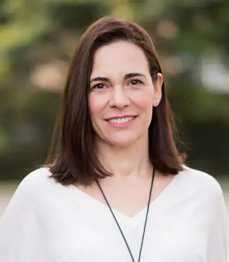 A woman with straight brown hair wearing a white top and a black necklace stands outdoors, smiling at the camera with a blurred green background.
