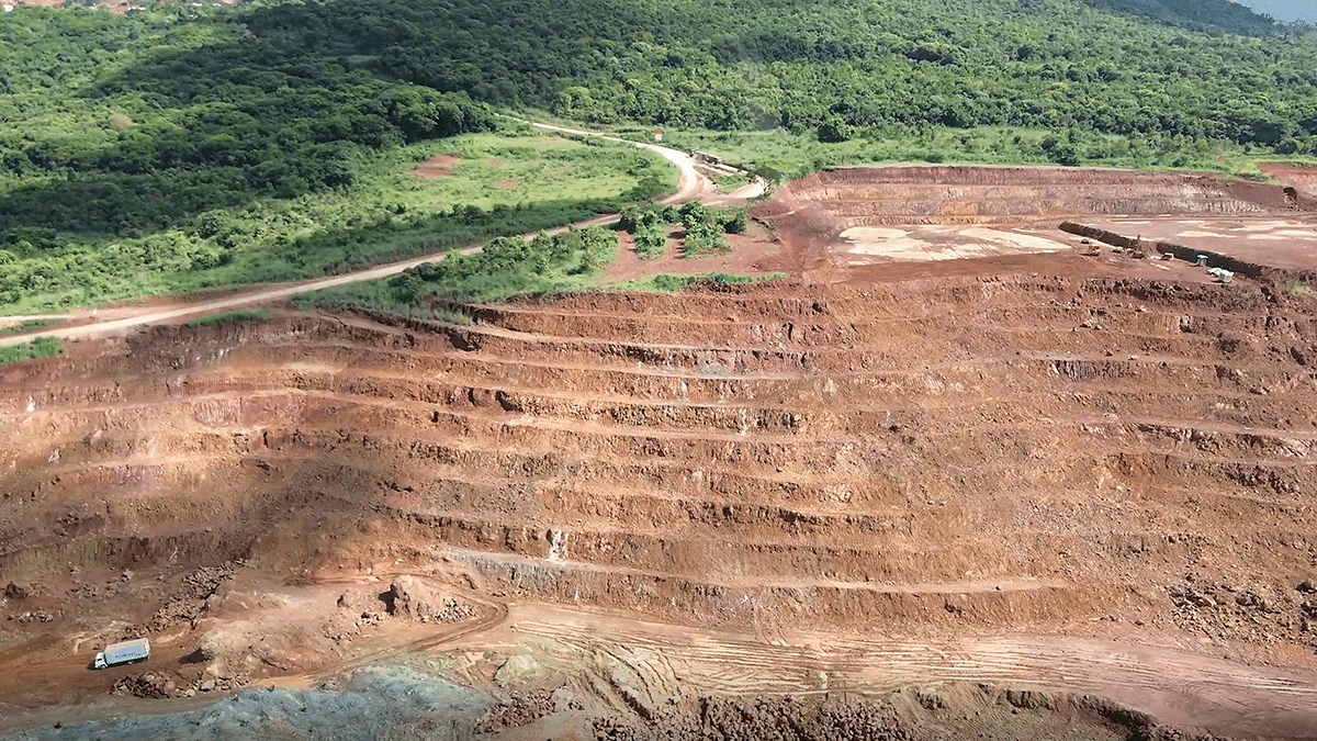 A terraced open-pit mine with reddish-brown soil is shown in the foreground; a green, forested area and a dirt road appear in the background.