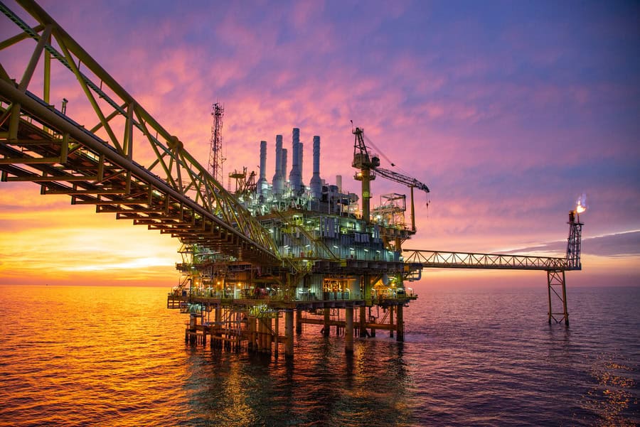 An offshore oil platform stands in the ocean at sunset, showcasing its infrastructure journey with industrial structures, cranes, and a gas flare silhouetted against a colorful sky.