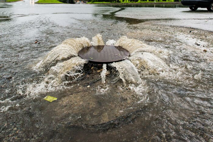 Water gushes out from beneath a manhole cover on a flooded street after heavy rainfall.