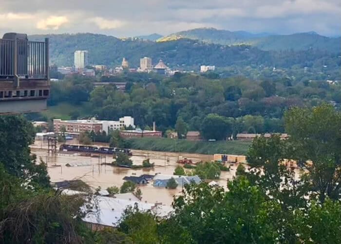 Aerial view of Asheville, NC with widespread flooding, submerged buildings, and surrounding hilly landscape under a cloudy sky.