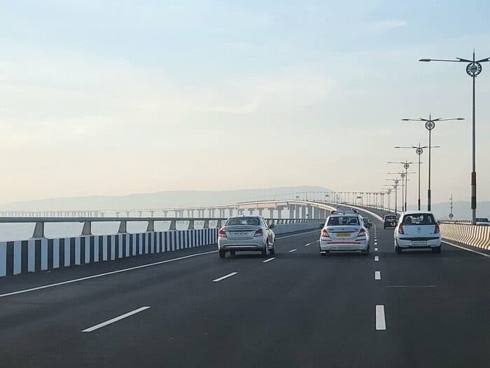 Four cars driving on a wide, multi-lane bridge during the day, with lamp posts lining the sides and distant hills visible in the background.