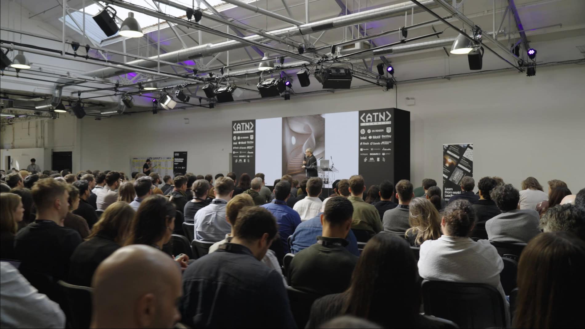 A large audience sits facing a stage where a speaker presents at a technology conference in a modern, industrial-style venue.