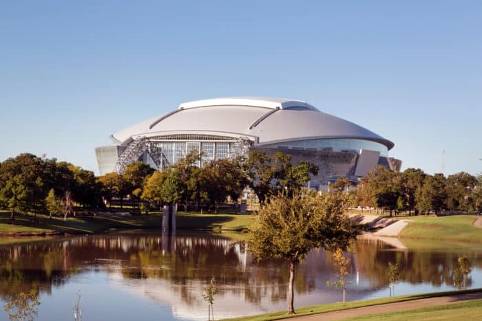 Large modern stadium with a curved roof surrounded by trees, reflected in a pond under a clear blue sky.