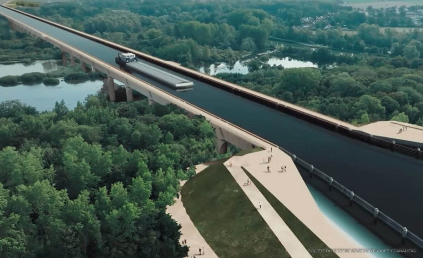 A cargo ship and a truck travel across a wide elevated aqueduct bridge spanning a forested area with people walking below.