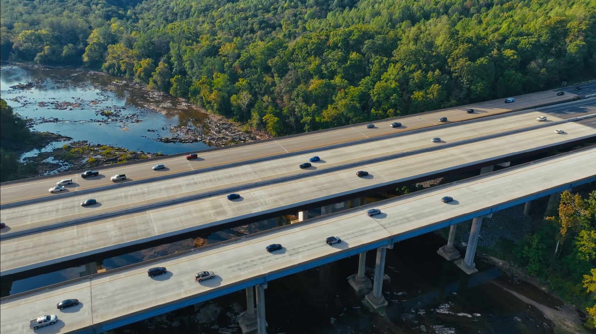 Aerial view of two parallel highways with cars driving, surrounded by trees and a river below.