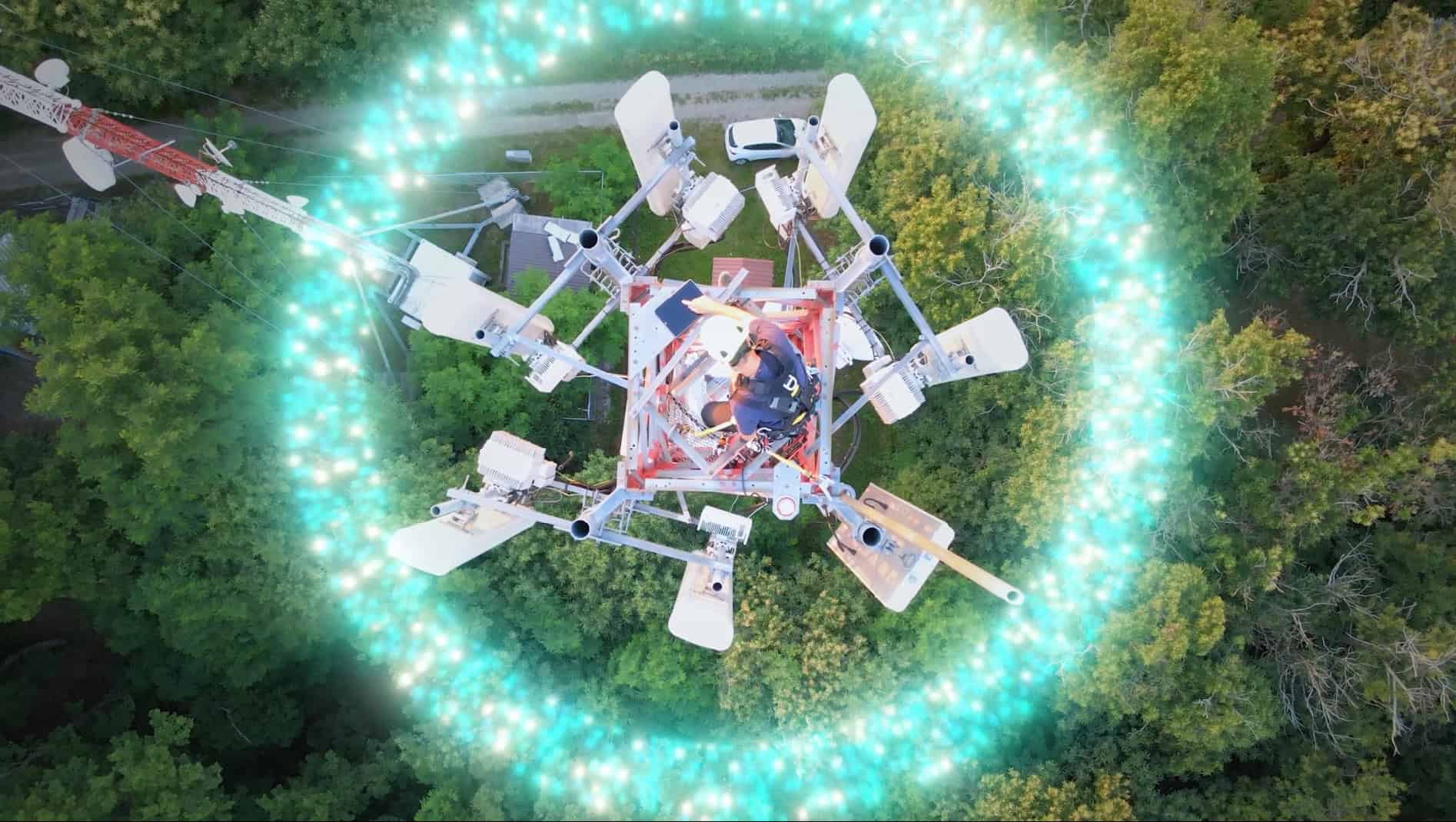 Aerial view of a cell tower surrounded by a glowing circular light effect, set in a forested area with a road and parked vehicles nearby.