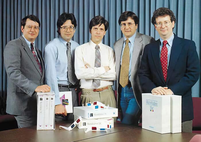 Five men in formal attire posing around a table with early software boxes of MicroStation in an office setting.