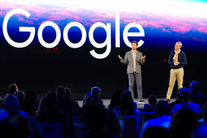 Two people stand on stage in front of a large screen displaying the word "Google," addressing an audience.