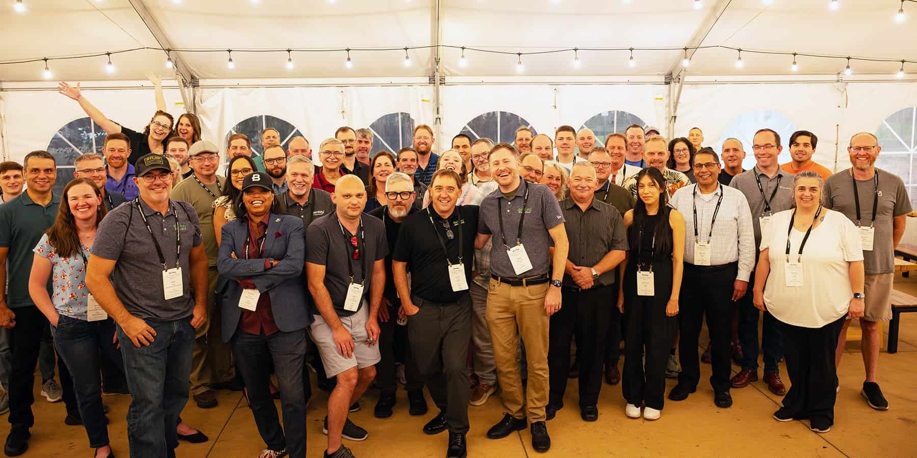 A large group of people pose and smile for a group photo inside a tent, many wearing name badges, under string lights at a Bentley Master Classes event.