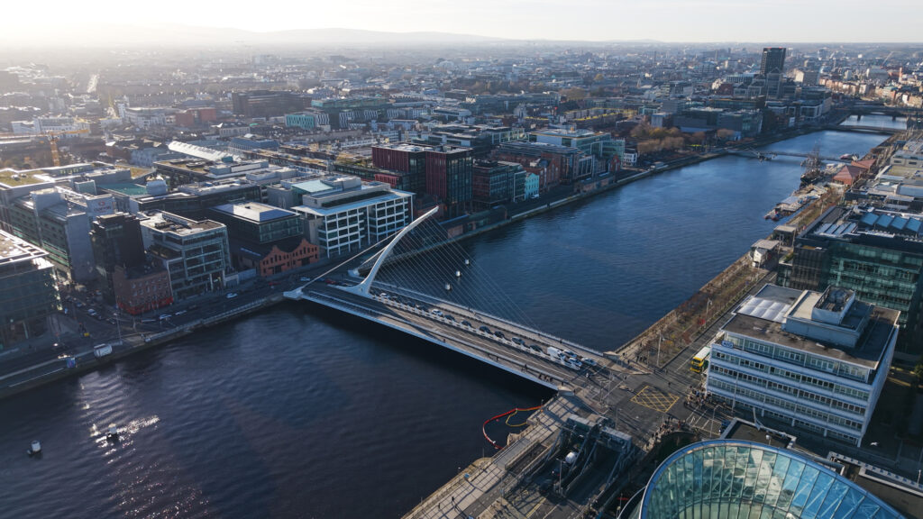 Aerial view of a modern bridge spanning a river in a city, with surrounding buildings and roads visible under a clear sky.