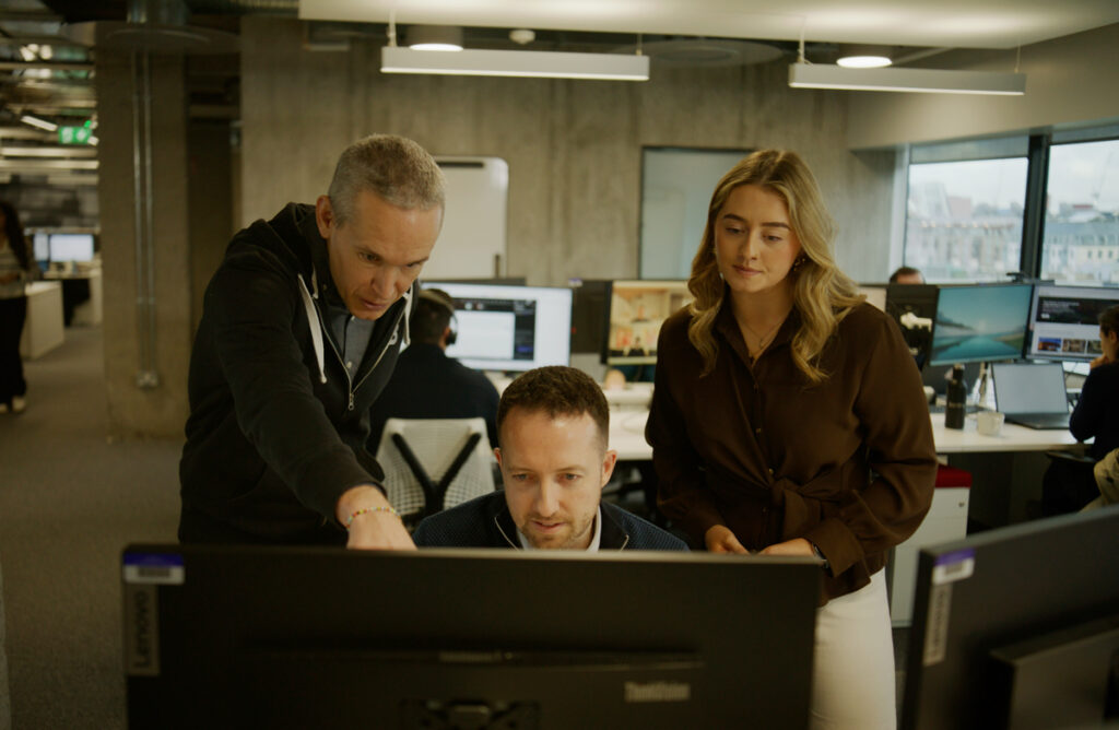 Three people in an office work together at a computer, with one person sitting and two standing as they look at the screen. Other office workers are visible in the background.