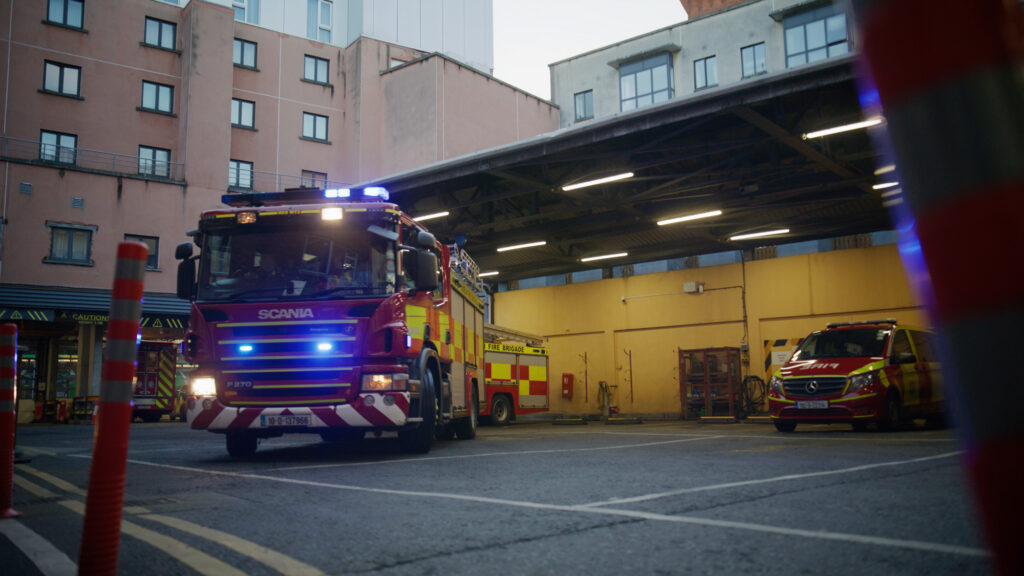 A fire truck with flashing lights exits a fire station garage in Dublin, while another fire truck and emergency vehicle are parked inside. Urban buildings surround the station.