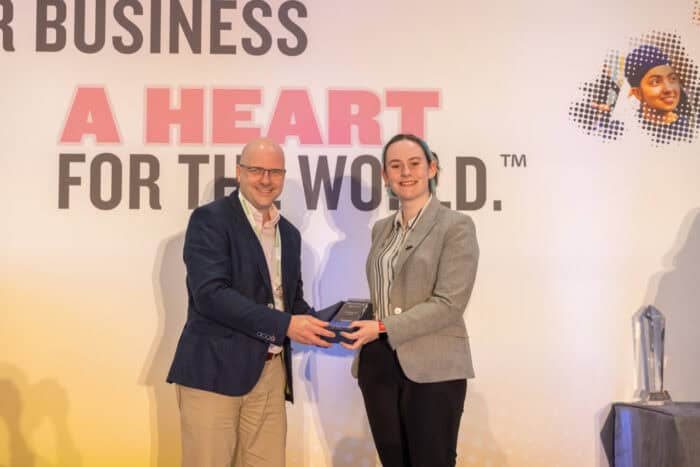 A man and a woman smiling, holding an award between them at a business conference with the slogan "a heart for the world" in the background.