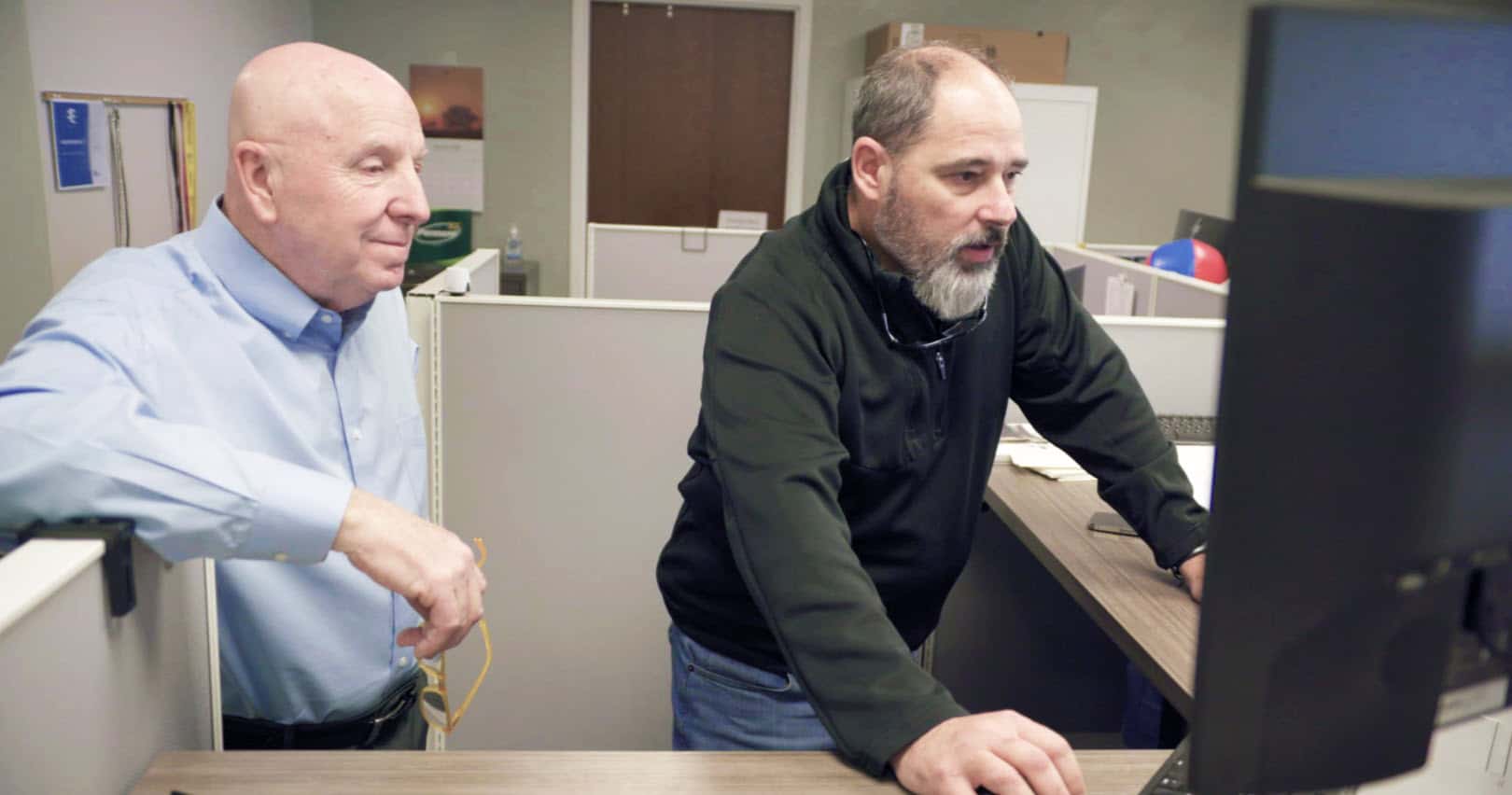 Two men stand at a desk in an office, looking at a computer monitor displaying OpenSite+; one types while the other observes, collaborating on site design.