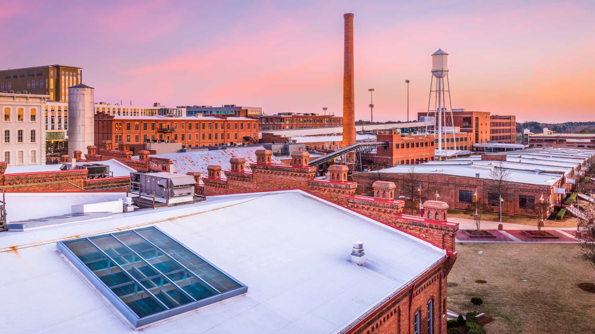 Red brick industrial buildings with a tall smokestack and a water tower, built to last, stand under a pink sunset sky in an urban area.