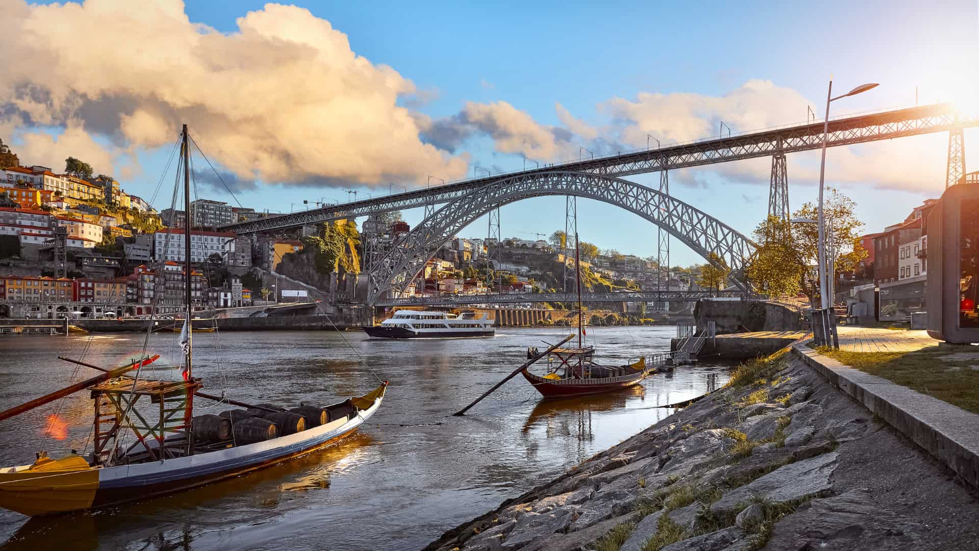A large arched steel bridge spans a river where boats are moored along the shore; city buildings from the Portuguese Water Utility climb the hillside, as sunlight breaks through clouds—a tranquil scene undisturbed by passing heat waves.