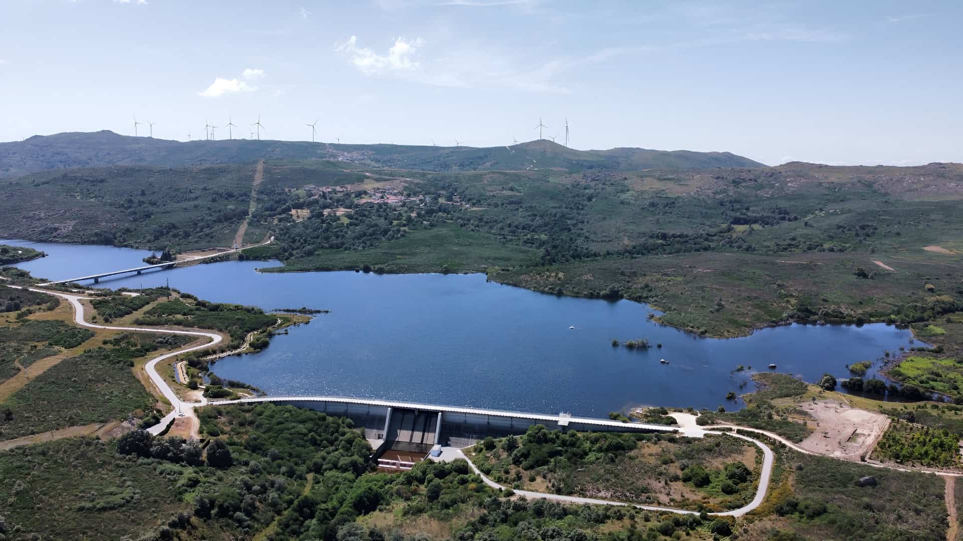Aerial view of a dam with a reservoir surrounded by hills and vegetation; wind turbines are visible on the ridge in the background, highlighting innovative software used by the Portuguese Water Utility to manage resources amid increasing heat waves.