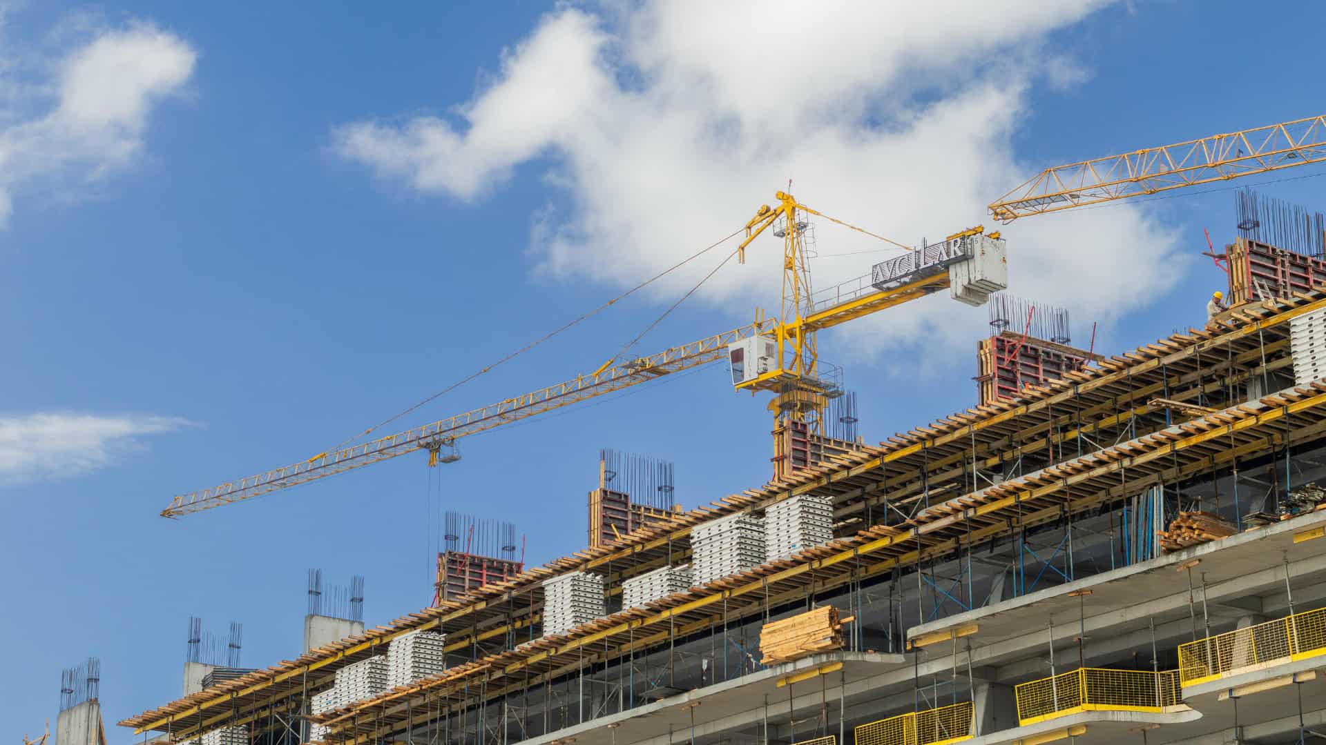 A yellow tower crane stands above a multi-story building under construction, illustrating how AI in infrastructure can help address staffing challenges amid scaffolding, concrete structures, and blue sky in the background.