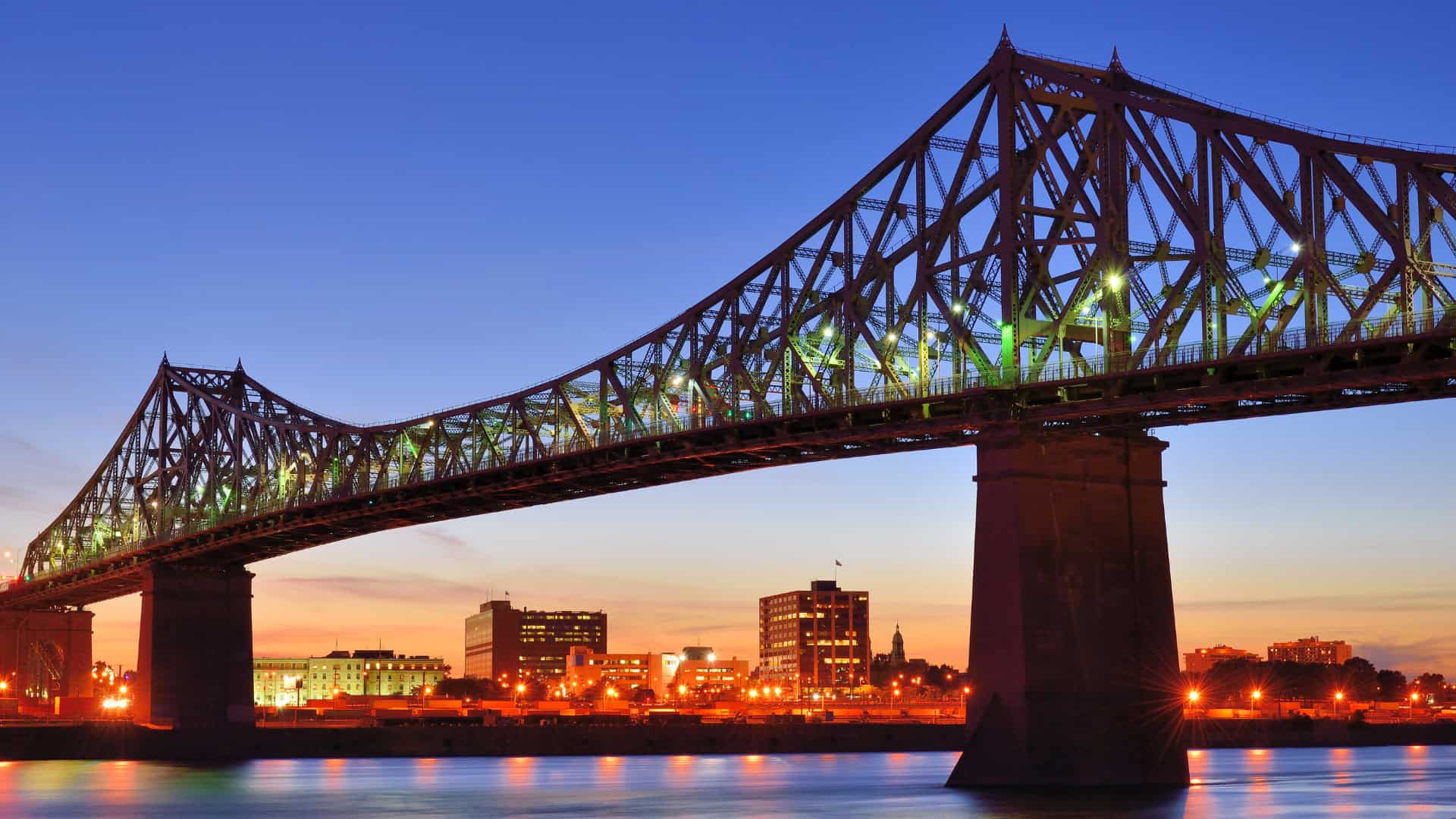 A large steel bridge, a testament to robust infrastructure, spans a river at sunset, with city buildings illuminated by lights in the background.