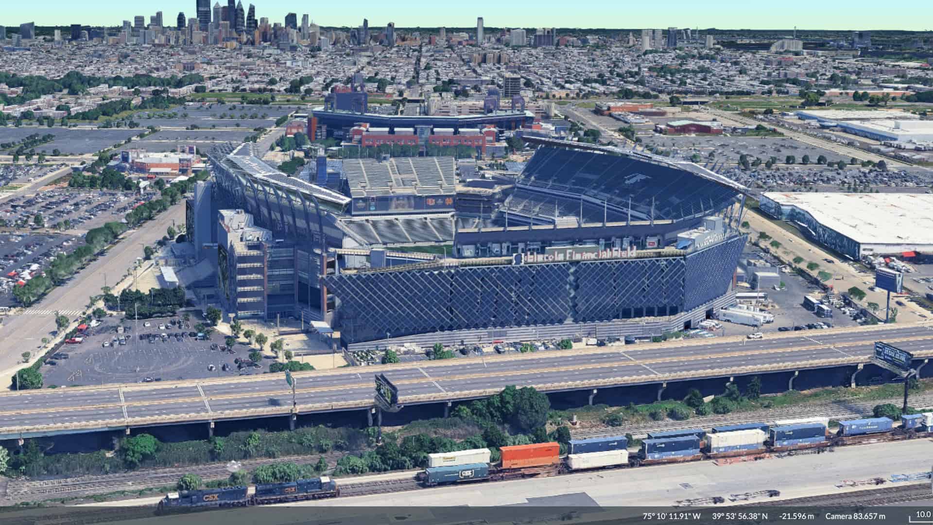 An aerial 3D map view of a large World Cup stadium near a highway with freight trains, surrounded by urban buildings and the bustling cityscape in the background.