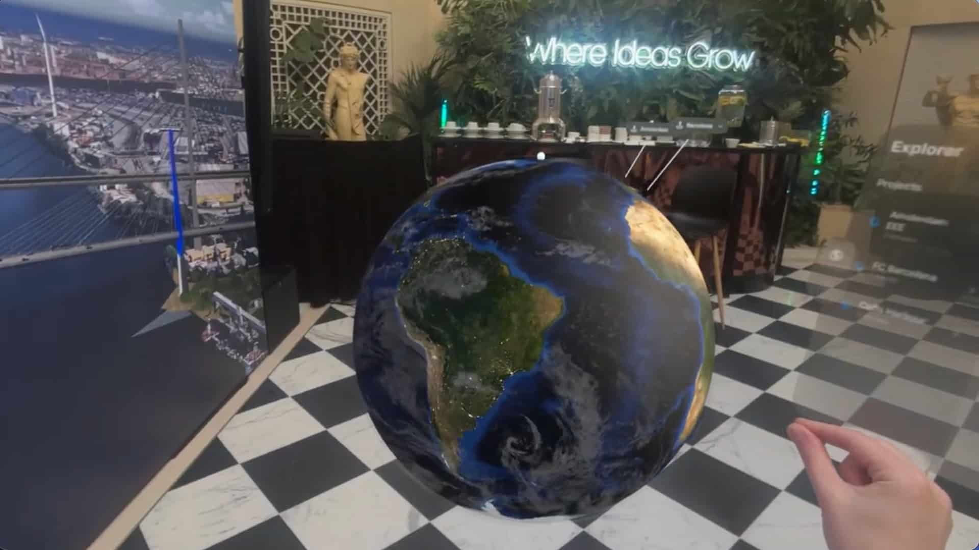 A person interacts with a virtual 3D Cesium globe in a room with checkered flooring, posters, plants, and a neon sign reading "where Ideas Grow.