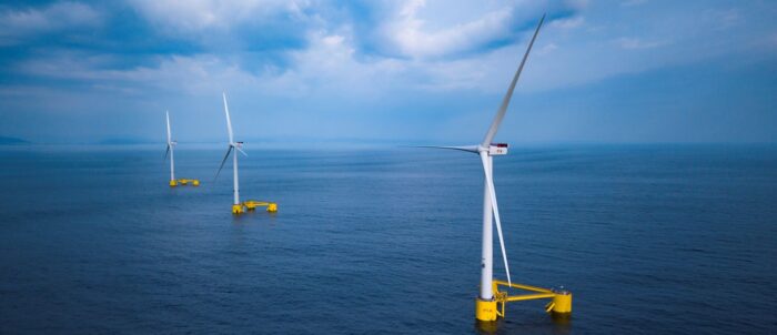 Three offshore wind turbines with yellow bases stand in a calm ocean, under a hazy blue sky.